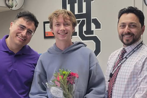 A man in a purple shirt, a teen boy in a hoodie holding flowers, and a man in a button-down shirt smile