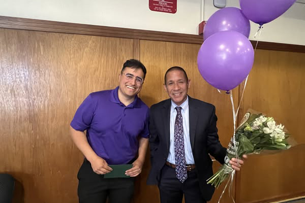A man in a purple shirt and a man in a suit holding purple balloons and flowers smile