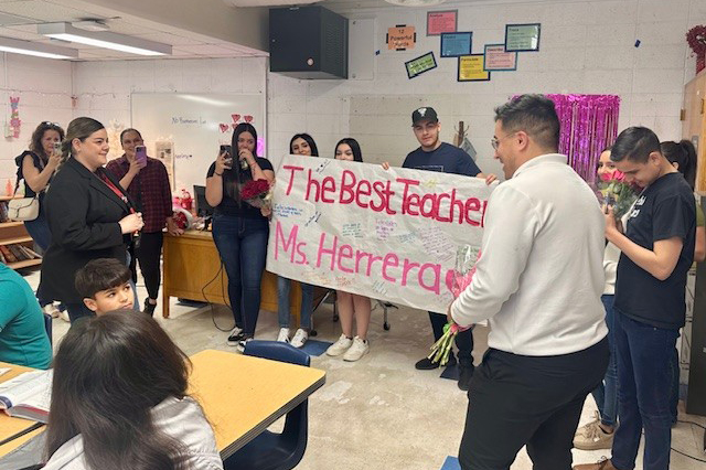 A woman smiles as her students hold up a banner reading The Best Teacher Ms. Herrera