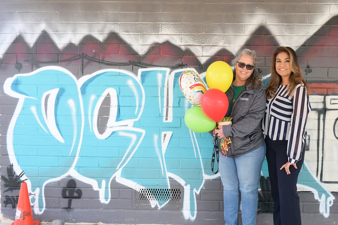 A woman holding a balloon bouquet and a woman in a striped shirt stand in front of an Ochoa graffiti sign