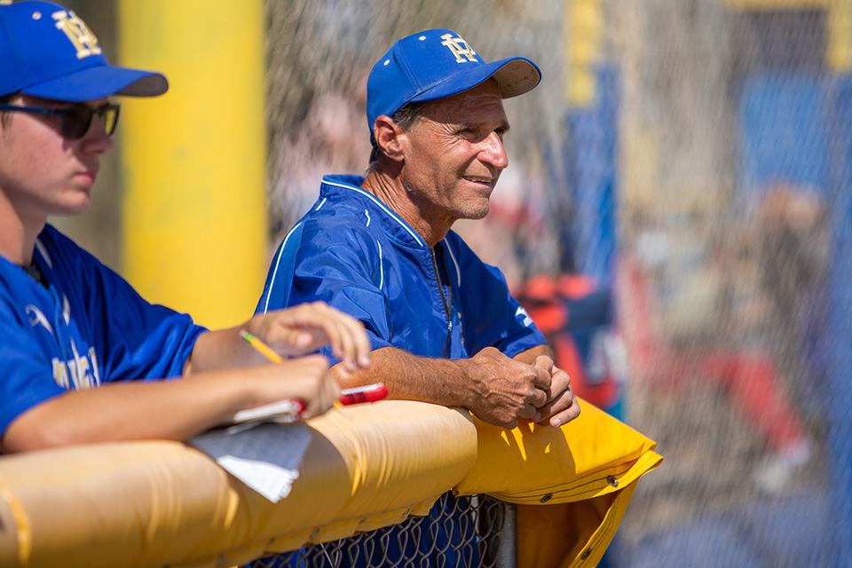A coach in a blue baseball hat and jersey stands on the sidelines next to a player