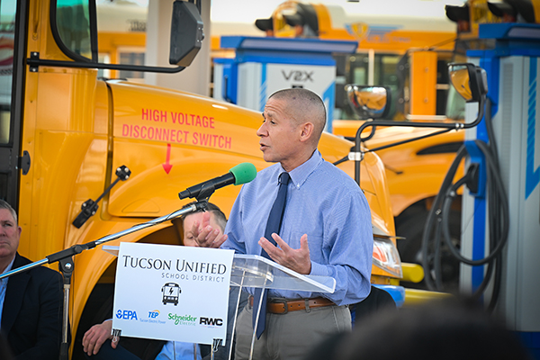 A man stands in front of an EV bus, addressing the audience