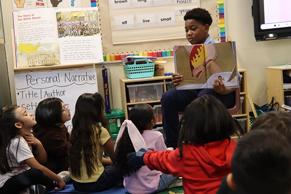 A boy reads to a group of younger students sitting on the floor