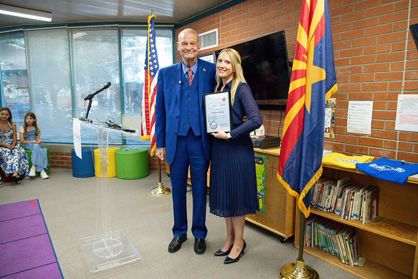 A man in a blue suit and woman in a blue dress pose for a photo, woman holds certificate