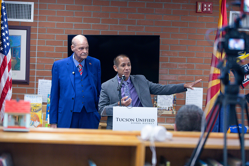 A man in a blue suit looks on as a man in a gray suit talks to the audience