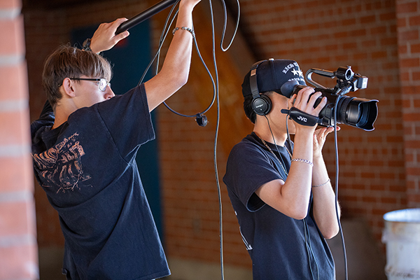 A teen boy holding a boom mic stands behind a teen boy holding a video camera