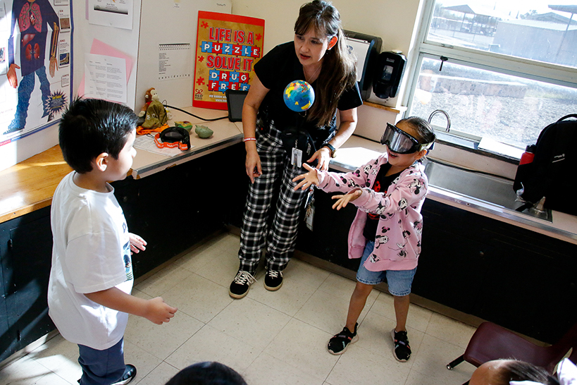 A little girl wearing impairment goggles throws a ball to a little boy