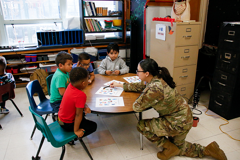 A woman in a military uniform crouches down to talk to kids at their table