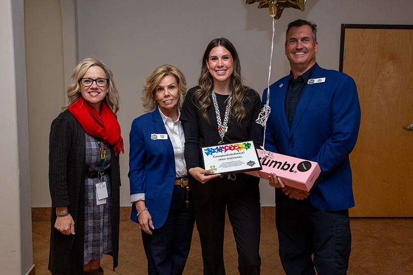 A woman, third from left, holds an award certificate with two women next to her and a man, right, holding a pink box of cookies and a balloon