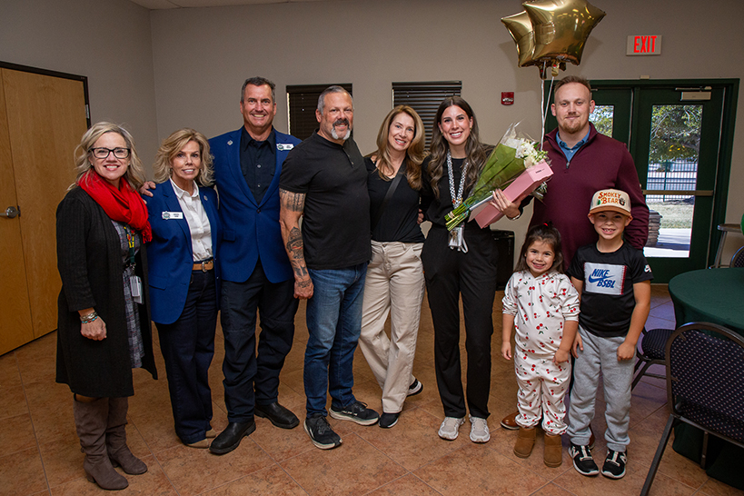 A group of people surround a woman holding flowers, an award certificate and a box of cookies