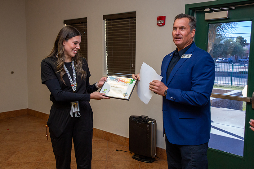 A woman smiles as she looks at her award certificate, next to a man in a blue suit