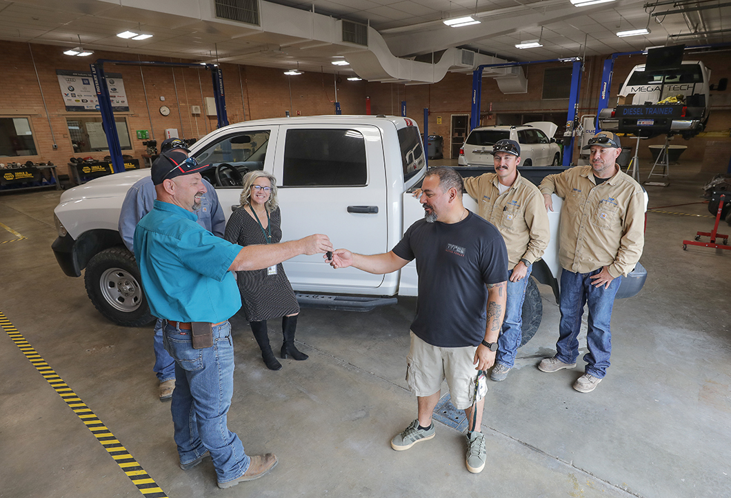 A man in a blue shirt hands the keys to a white pickup truck to a man in a black shirt, while a woman in a gray dress and two men in tan shirts look on