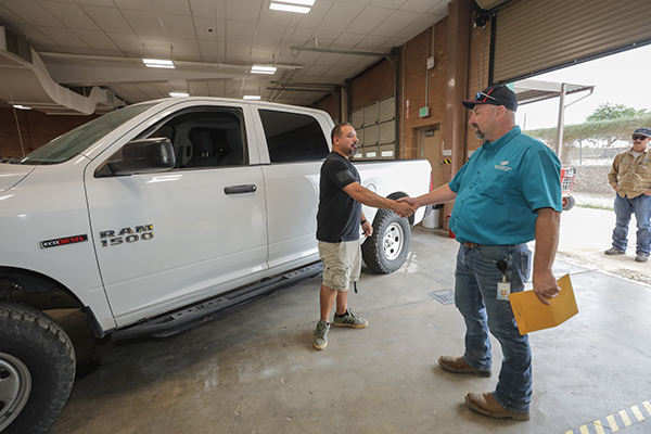 A man in a black shirt, standing in front of a white pickup truck, shakes a man in a blue shirt's hand