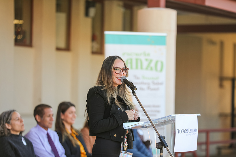 A woman in glasses and a black suit talks into a mic