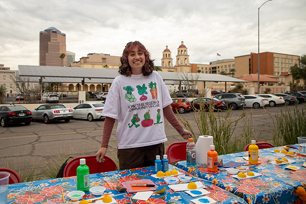 A woman wearing a shirt with veggies on it smiles as she stands behind a table