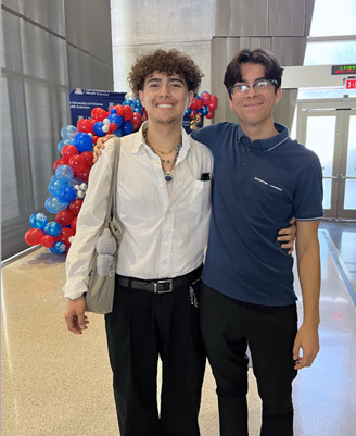 Two teen boys smile in the entryway of a building, with a balloon arch behind them
