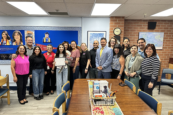 A teen girl, center left, holds an award certificate surrounded by family and teachers in a classroom