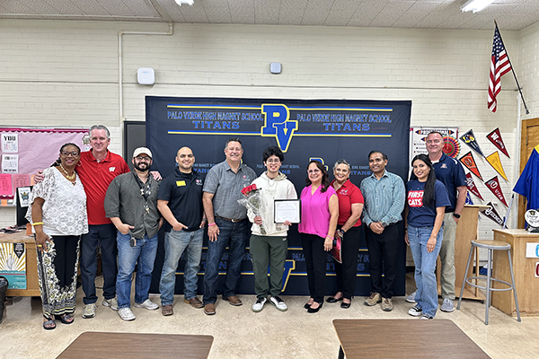 A teen boy, center, holds an award certificate surrounded by family and teachers in a classroom