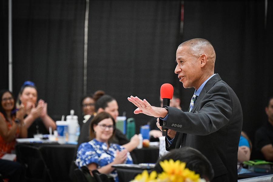 A man in a suit speaks in a red microphone, addressing a crowd of administrators