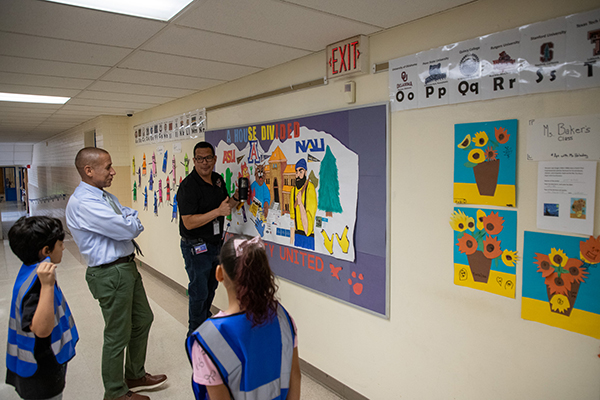 A man in a black outfit and glasses shows a man in a blue shirt and green pants and two students in blue vests a wall of student artwork