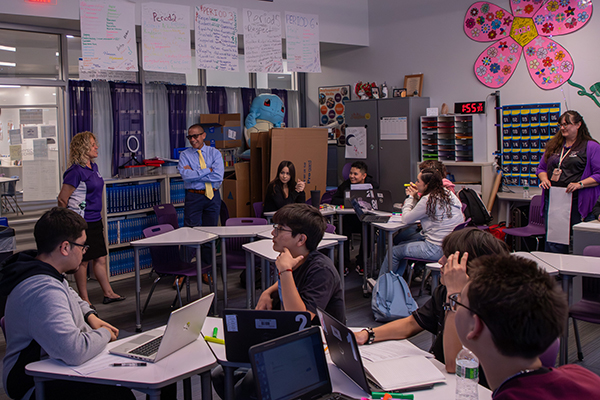 A man in a blue shirt, navy pants and yellow tie talks to a woman in a purple shirt in the back of a classroom of high school students