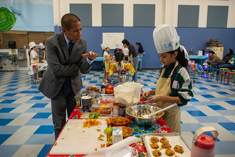A man in a suit talks to a girl in a chef's hat who's cooking up some treats