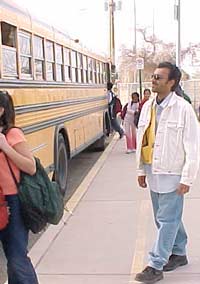 Students stand outside at a school bus stop waiting to board.